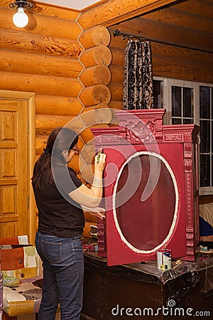 Old Lady in glasses with brush aging wooden cupboard door with red carved ornaments in wooden house. Meticulous process Stock Photo