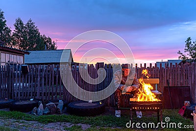 An old lonely man sits sadly on a bench at a village house behind a fence looking into the fire of a barbecue fire Stock Photo