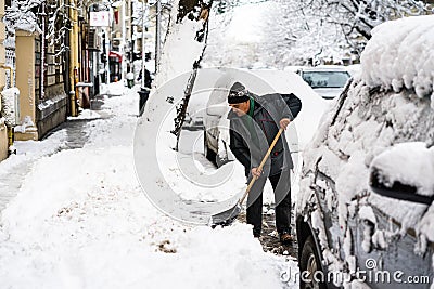 Old man, male in winter coat cleaning, shoveling driveway, street from covered falling snow with shovel, residential houses, cars Editorial Stock Photo