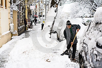 Old man, male in winter coat cleaning, shoveling driveway, street from covered falling snow with shovel, residential houses, cars Editorial Stock Photo