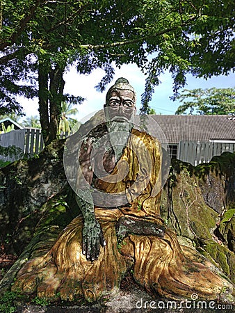 old man statue, faded, faded paint. wearing a yellow shirt, sitting. Stock Photo