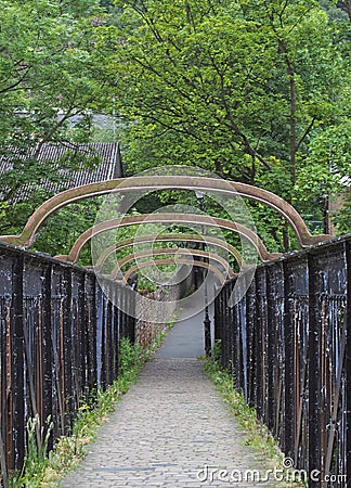 Old metal pedestrian footbridge crossing a railway line leading to a narrow alley surrounded by trees and rustic buildings Stock Photo