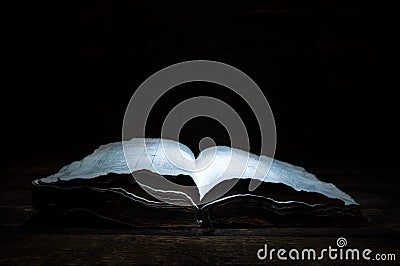 An old open book is lying on a wooden table in the dark. A light shines on the book from above. Holy Bible. Stock Photo