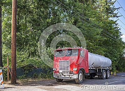 Old red big rig cabover semi truck with tank trailer standing on the road shoulder on the green trees background Stock Photo