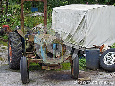 Old rusting abandoned tractor next to a dirty tarpaulin covered trailer and junk in a farmyard Stock Photo