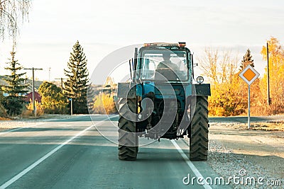 Old rusty vintage tractor of blue color is driving along the road in a village in Russia in summer, rural life in the Saratov Editorial Stock Photo