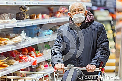 Old senior european man wearing protective facial mask pushing shopping cart in the supermarket. Shopping during COVID-19 concept Stock Photo