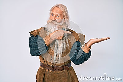 Old senior man with grey hair and long beard wearing viking traditional costume amazed and smiling to the camera while presenting Stock Photo