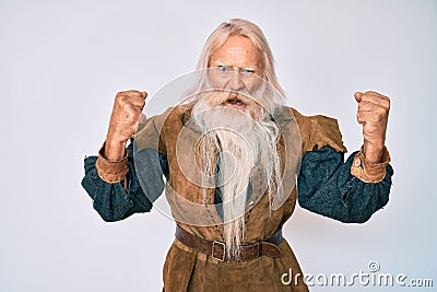 Old senior man with grey hair and long beard wearing viking traditional costume angry and mad raising fists frustrated and furious Stock Photo