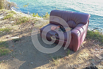 Old torn sofa with burgundy color. Red ruined couch on sea shore. Hole in seat and torn foam. Abstract composition of old couch Stock Photo