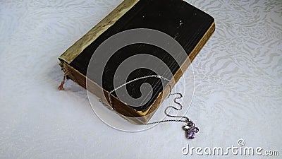 An old vintage Bible and a silver crucifix on a chain with purple crystals on a white tablecloth. Stock Photo