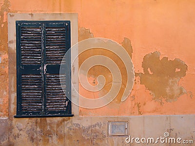 Old window with green painted closed wooden shutters on an orange Mediterranean ocher colored orange cracked peeling flaking wa Stock Photo
