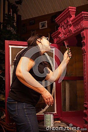 Old woman in glasses with big brush in hand aging wooden cupboard with bright red carved ornaments. Meticulous process Stock Photo