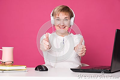 Old woman in her 60s works on computer, wearing headphohes. Laptop on white table and Pink background. She make thumbs up gesture Stock Photo