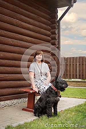 An elderly woman is sitting on a village bench with a black schnauzer near a wooden house in the countryside Stock Photo