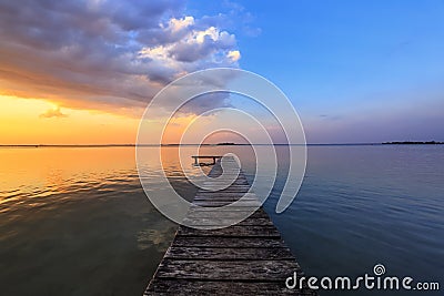 Old wooden jetty, pier reveals views of the beautiful lake, blue sky with cloud. Sunrise enlightens the horizon with orange warm Stock Photo