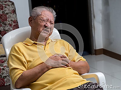 Older Asian men sitting on a chair at living room with heart attacks.Both old man`s hands on breast because of hard breathing Stock Photo