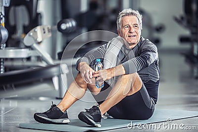 Older male rests with a bottle of water sitting on a mat during a gym workout Stock Photo