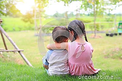 Older sister hugs little brother by the neck, shoulders sitting on green grass field. Two adorable Asian children sitting and Stock Photo