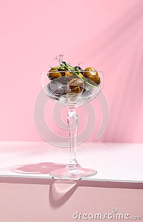 Olive Kalamata in cocktail glass. Black and green olives food on white table with pink wall. Day sunlight with hard shadow of fern Stock Photo