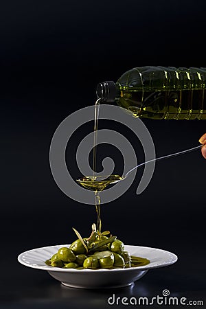 Olive oil being poured from a plastic bottle into a spoon and falling into a plate with olives Stock Photo