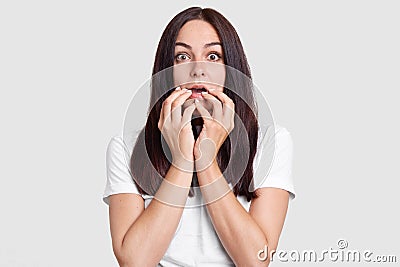 Omg, its terrible! Shocked worried woman with scared facial expression, keeps hands near mouth, hears shocking news, wears white c Stock Photo