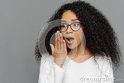 Omg, no way. Scared emotional young dark skinned woman covers opened mouth, feels impressed, has curly hair, wears transparent Stock Photo
