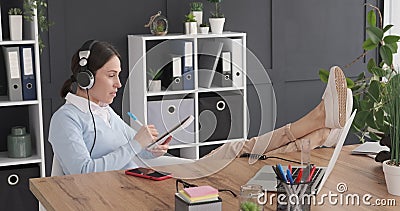 Onderneemster met voeten omhoog op bureau terwijl het luisteren muziek en het maken om lijst te doen stock footage Onderneemster met voeten omhoog op bureau terwijl het luisteren muziek en het maken om lijst te doen stock footage