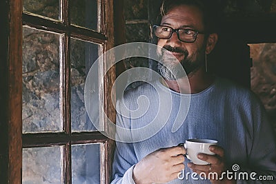 One mature man at home smile and enjoy indoor relax drinking tea or coffee and looking outside the window. People in winter chalet Stock Photo