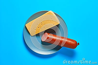 One piece of cheese in form of triangle near half of long sausage on gray round plate lies on blue desk on kitchen or market. Top Stock Photo