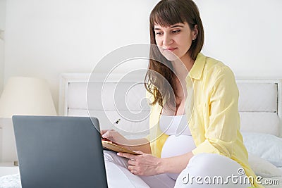 One pregnant european woman working from her home while she is sitting on a white bed at day time Stock Photo