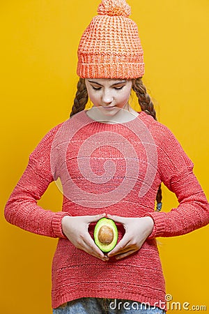 Human Health Concepts. Teenager Girl In Coral Knitted Clothing With Split Avocado Fruit In Front of Belly as a Demonstration of Stock Photo