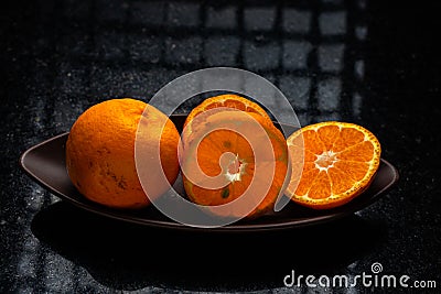 One whole tangerine and one cut in a brown canoe-shaped dish on a black stone table with a window reflection Stock Photo