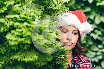 Ð¡onfused face woman in a santa claus red hat looks away through green fir tree branches in the forest. Christmas coming, sale Stock Photo
