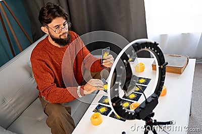 Online astrology. Male fortune teller is use a smartphone and ring light, reads tarot cards for predictions, telling Stock Photo