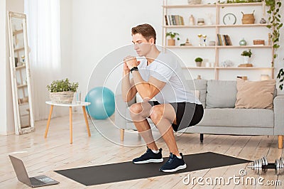 Online coach for home workout. Handsome man with fitness tracker, squats and looks at laptop Stock Photo