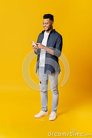 Online communication. Full length portrait of indian man using smartphone standing isolated on yellow background Stock Photo