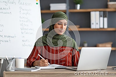 Islamic teacher in headscarf sitting at desk looking at laptop Stock Photo