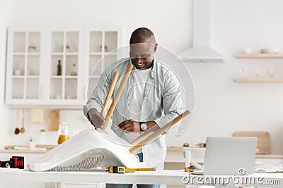 Online instructions of furniture assembly. Happy black handyman screwing detail to chair, using laptop computer Stock Photo