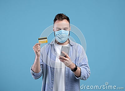 Online shopping during covid. Young guy in face mask purchasing goods on web, holding cellphone and credit card Stock Photo