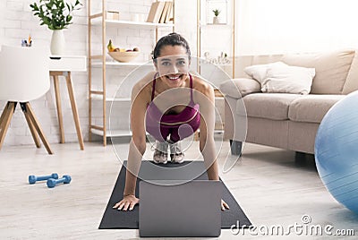 Online sports tutorials. Cheerful Latin girl doing plank exercise near laptop at home Stock Photo
