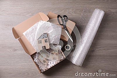 Open box with decorative house figures, adhesive tape, scissors, foam peanuts and bubble wrap on wooden table, flat lay Stock Photo