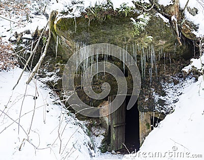 Open dark cellar door in sandstone rock cave with moss and lichens and long hanging icicles, white snow background. Stock Photo