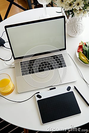 Opened laptop, graphics tablet and breakfast with juice on white table, empty screen for mockup design. Stock Photo