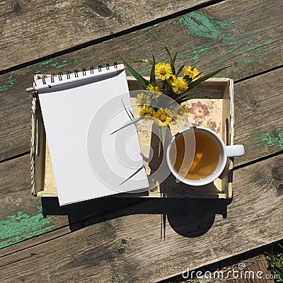 Opened notepad, cup of tea and small bouquet of wild flowers on a tray on a old wooden background. Stock Photo