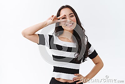 Optimistic cheerful young asian woman in striped t-shirt staying positive, show peace or victory sign over eye, hold Stock Photo