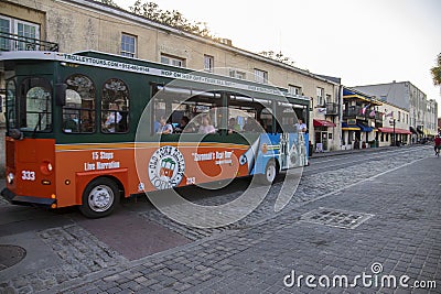 An orange and green trolley bus from Old Town Trolley driving on River Street with shops and restaurants and people walking Editorial Stock Photo