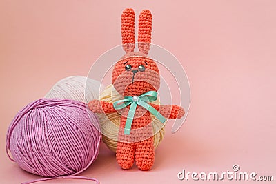 Orange knitted hare stands on the background of multi-colored balls of yarn on a pink background. There is a place for text Stock Photo