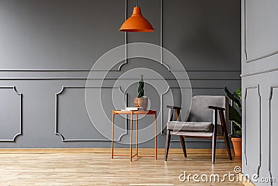 Orange lamp above table with plant next to armchair in grey apartment interior. Real photo Stock Photo