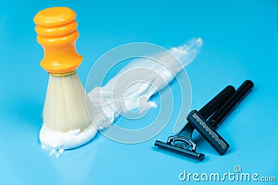 An orange shaving brush stands in whipping foam and two black razors on a blue background Stock Photo